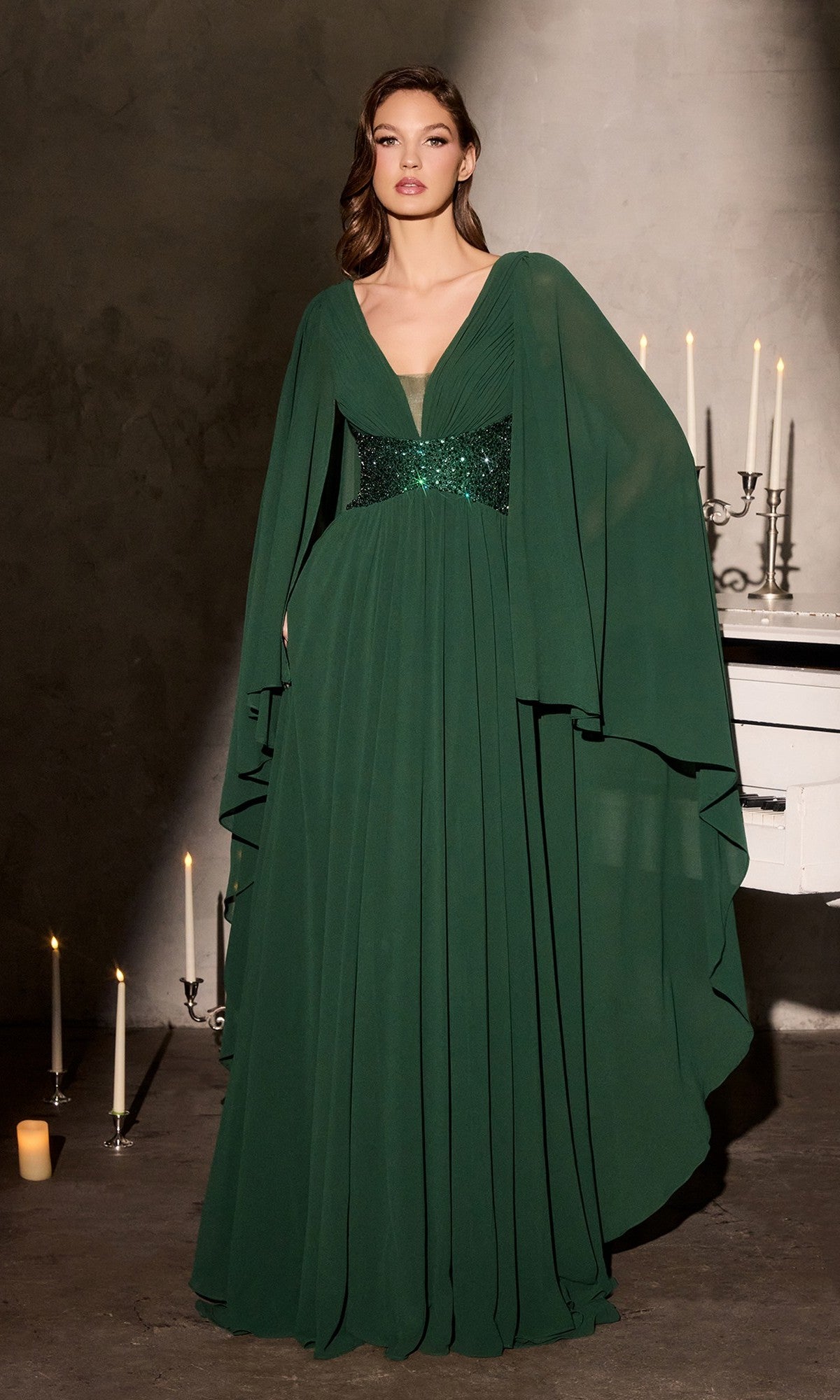 A woman stands indoors wearing the emerald Long Formal Dress: Ladivine CM8244, featuring cape-style sleeves and a beaded waistline; softly glowing candles illuminate the background.